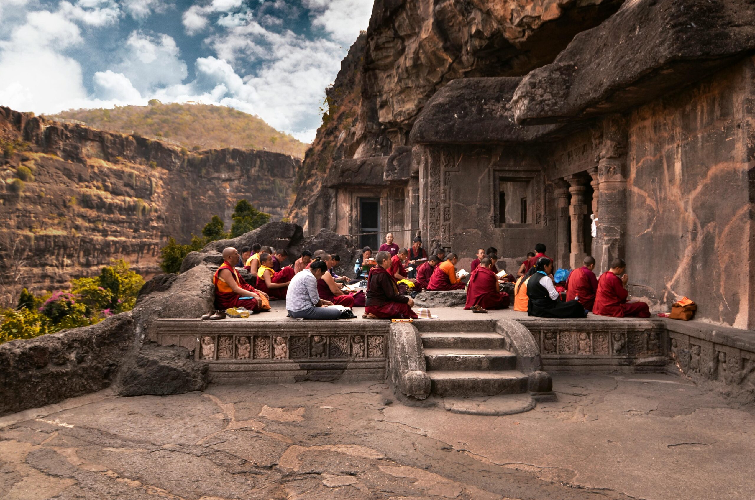 Group of monks in traditional robes meditating at an ancient rock temple during the day.